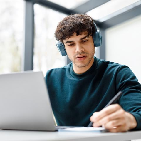 Junger Mann sitzt vor Laptop, trägt Kopfhörer und lernt