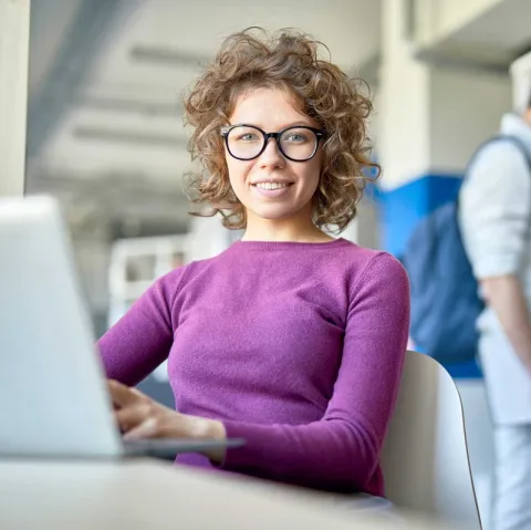 Symbolbild mit junge Frau, die vor dem Laptop sitzt und in die Kamera schaut