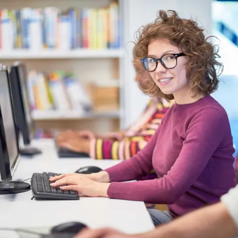 Studentin sitzt vor PC und tippt auf Tastatur. Im Hintergrund stehen Bücherragale.