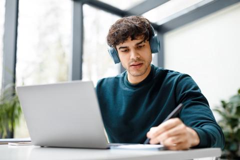 junger Mann mit Kopfhörern sitzt konzentriert vor Laptop und notiert sich etwas in ein Heft.