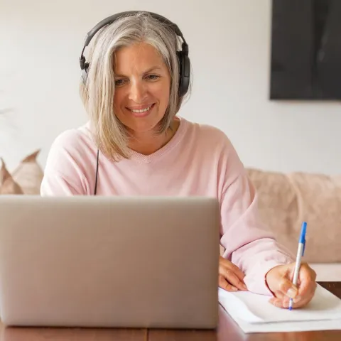 Frau, mittleren Alters, sitzt vor einem Laptop und schreibt auf ein Blatt.