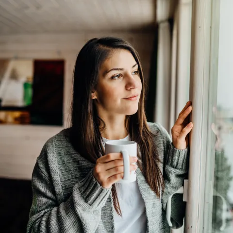 Junge Frau schaut aus dem Fenster und hält eine Tasse Tee in der Hand.