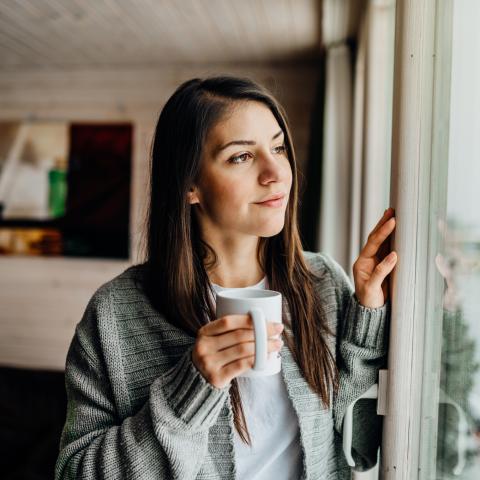 Junge Frau schaut aus dem Fenster und hält eine Tasse Tee in der Hand. 