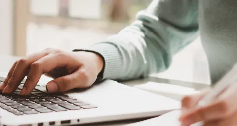 Es ist eine Tastatur eines Laptops zu sehen. Unscharf ein Person, die einen Stift in der Hand hält.