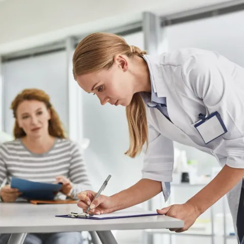 eine Medizinsche Praxisasstentin schreibt etwas in ein Buch. Eine Patientin sitzt im Hintergrund.