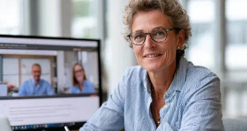 Frau, ca. 50 Jahre sitzt an PC und blickt in Kamera. Im Hintergrund ist ein Bildschirm mit Online-Teilnehmern zu sehen